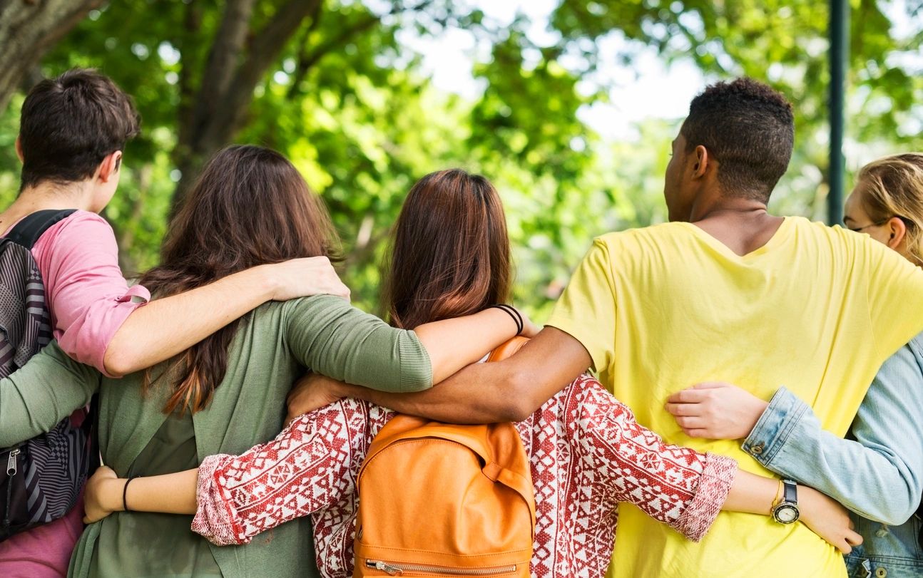 Four young people standing outdoors with their backs to the camera, arms around each other's shoulders, surrounded by green trees.