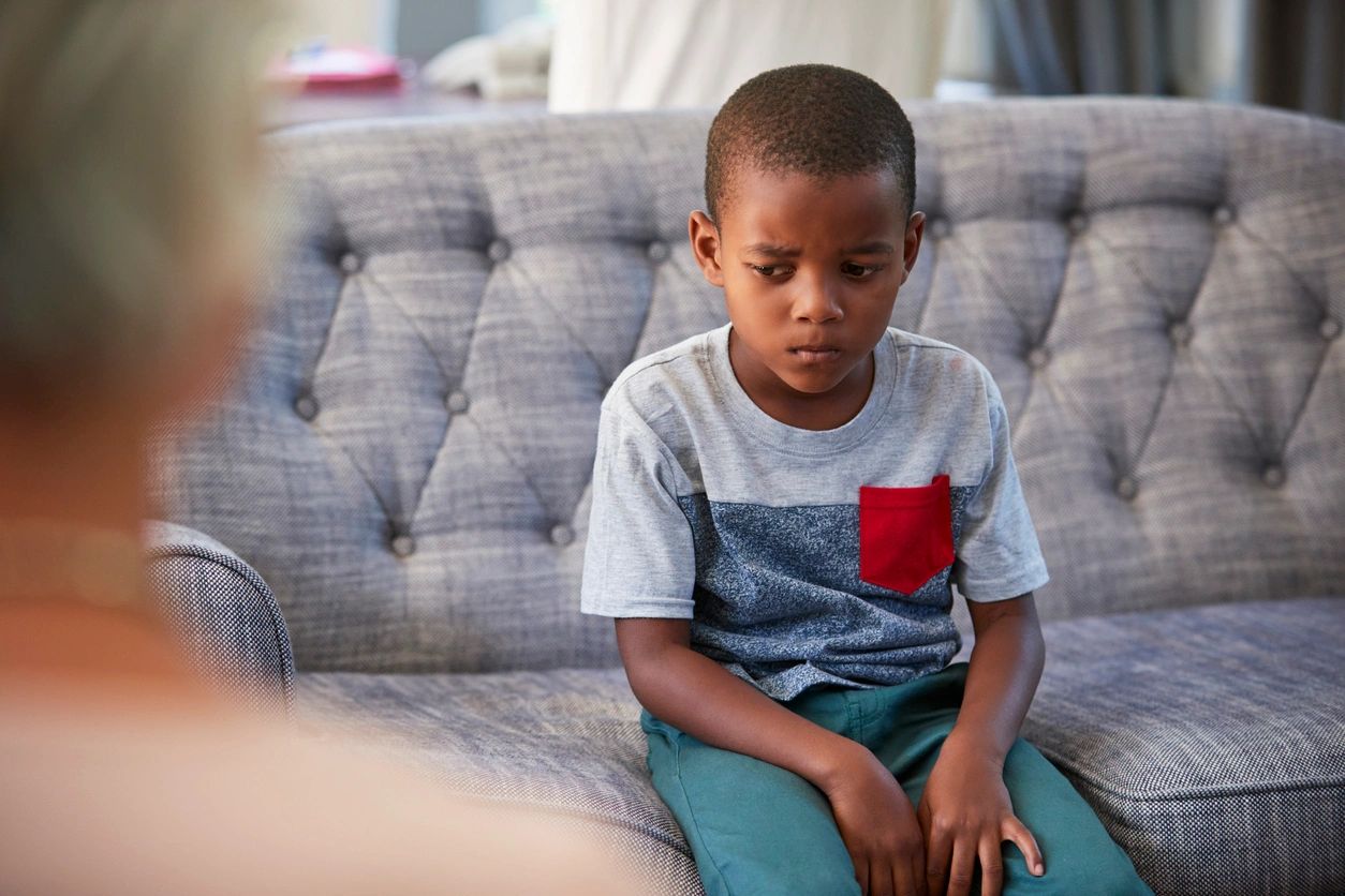 A young boy sits on a gray sofa, looking down with a serious expression, wearing a gray shirt with a red pocket and green pants.