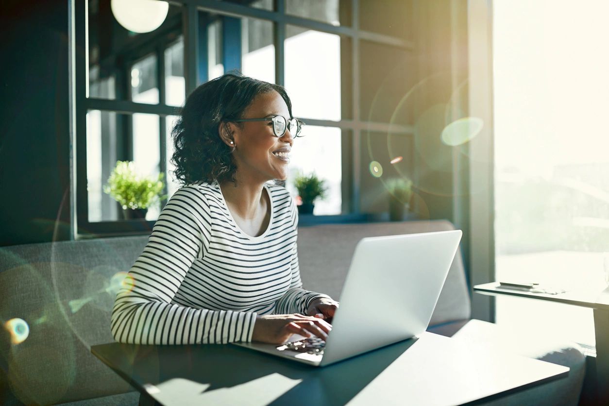 A person wearing glasses and a striped shirt is sitting at a table, smiling while working on a laptop. Sunlight streams through the window behind them.