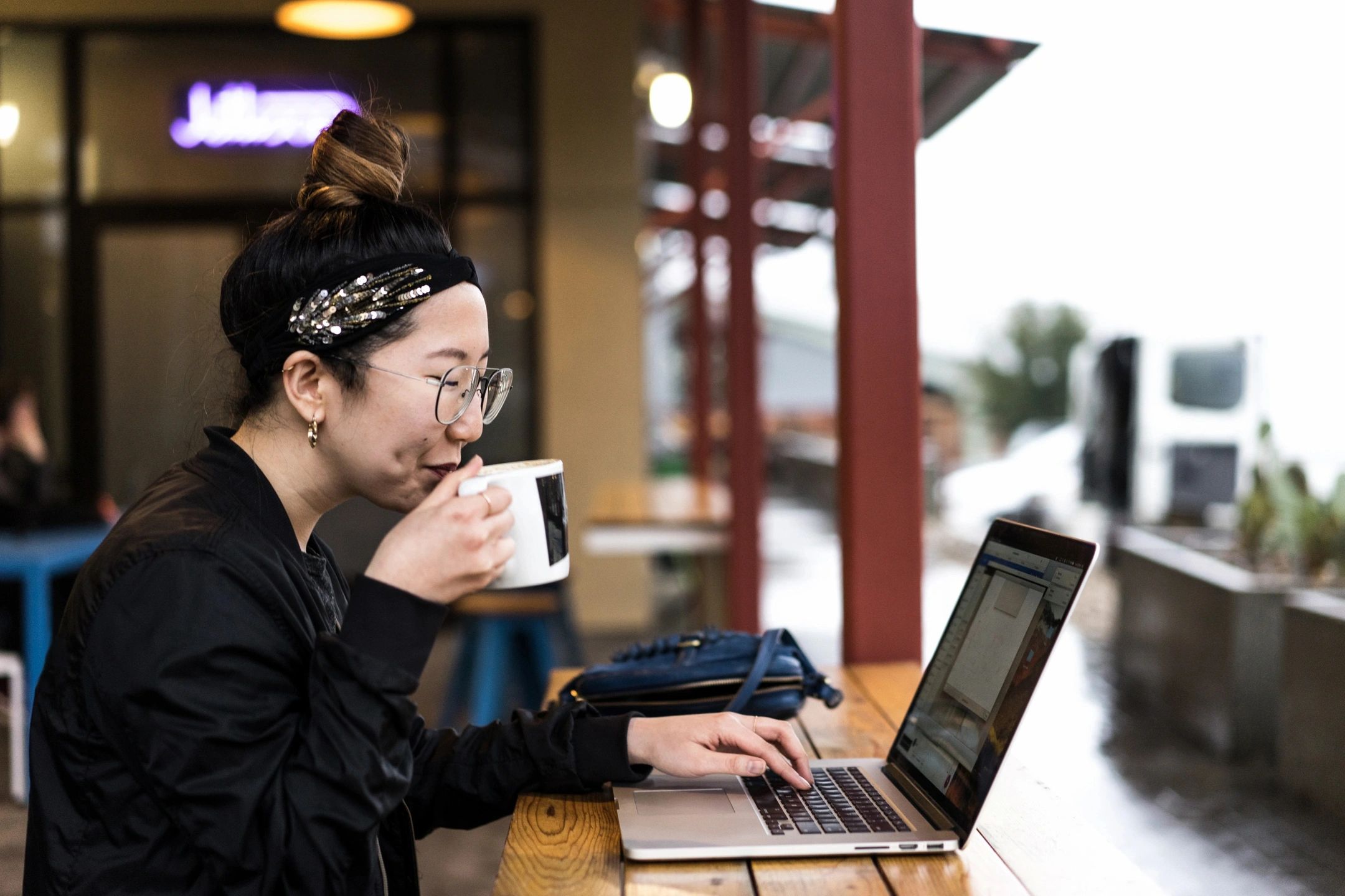 A person wearing glasses and a headband sips from a cup while using a laptop.