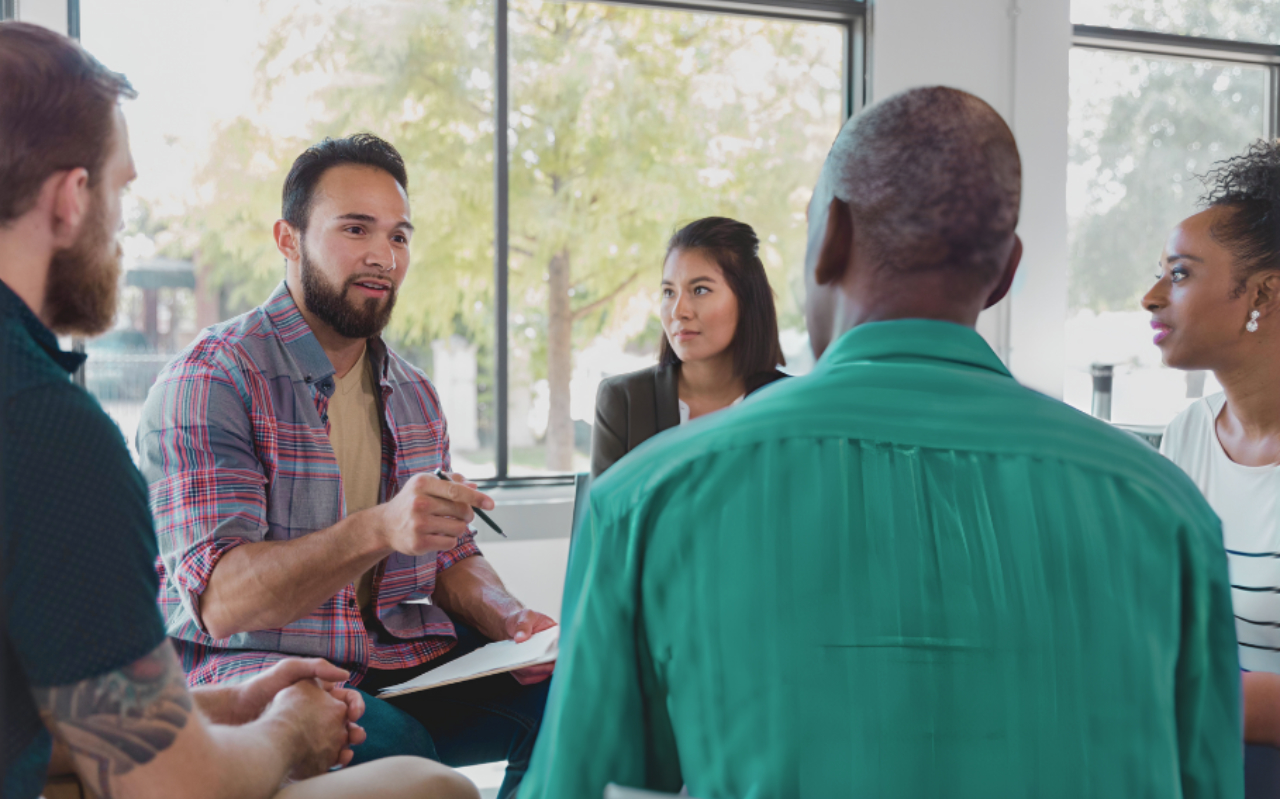 A man holding a notebook and pen speaks to a small group. Another man and a woman listen attentively in a brightly lit room with a window in the background.