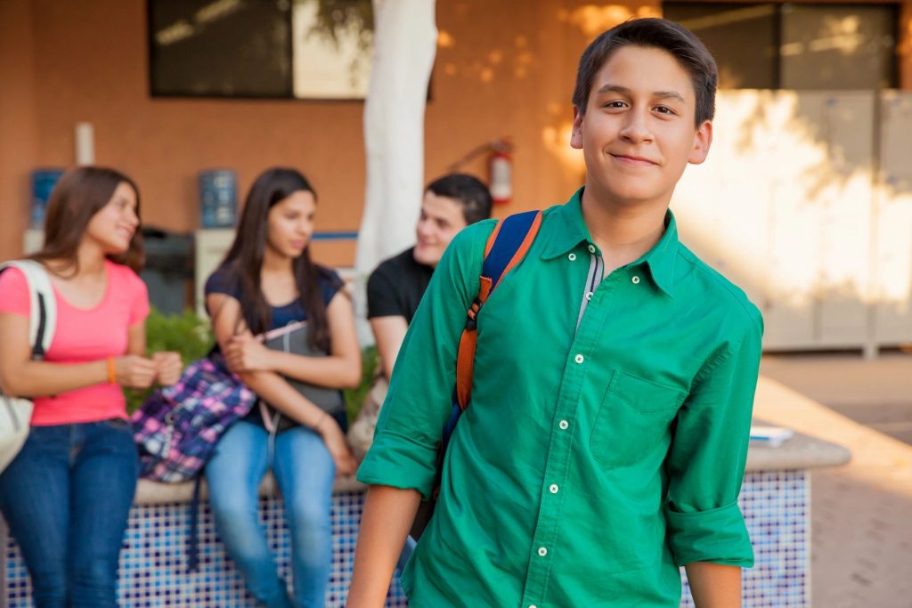 Teenage boy wearing a green shirt and backpack smiles at the camera while standing outside with three peers in the background.