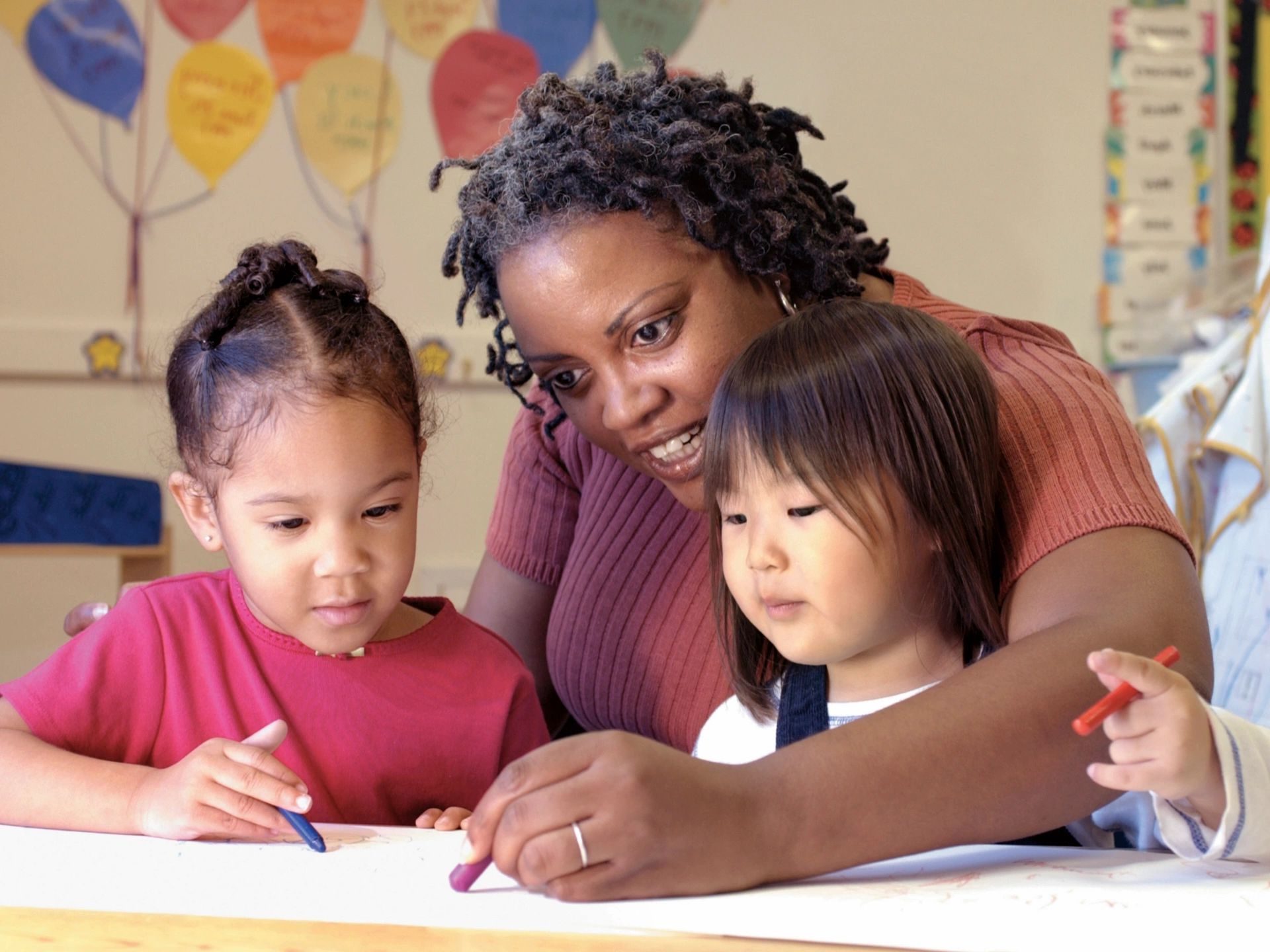 An adult helps two young children with coloring activities at a table in a classroom setting.