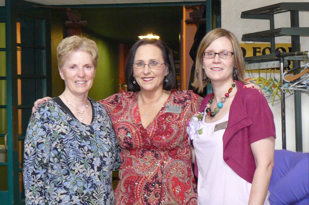 Three women involved with ISAS pose together indoors, smiling at the camera. They are wearing patterned clothes and name tags. Marcie Sheridan on on the far right.