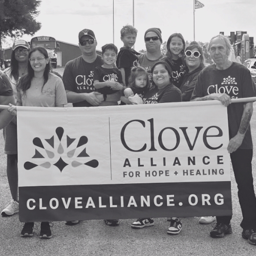 A group of people wearing "Clove Alliance" shirts, holding a banner that reads "Clove Alliance: For Hope + Healing, clovealliance.org.