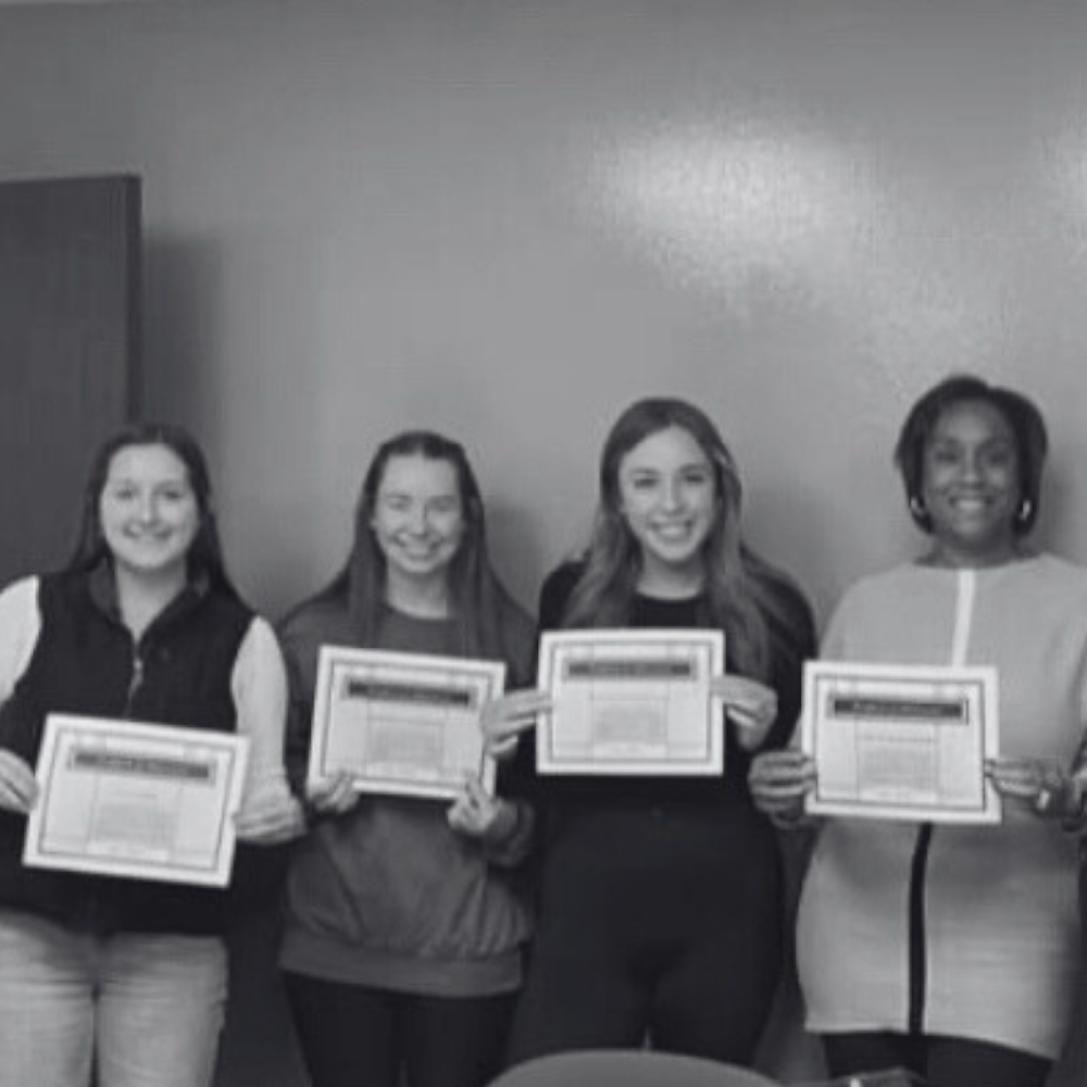 A group of four people stand indoors, holding certificates. The image is in black and white.