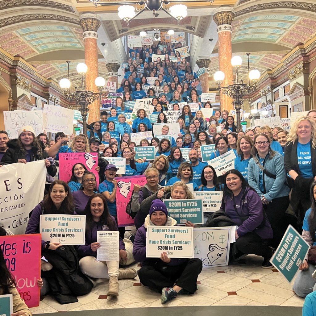 A large crowd gathers at the Illinois Capitol Building holding signs in support of funding sexual assault survivor services.