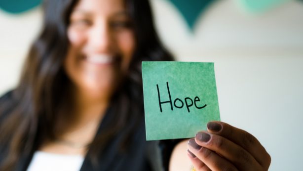 A person holds a green note with "Hope" written on it, smiling in the background.