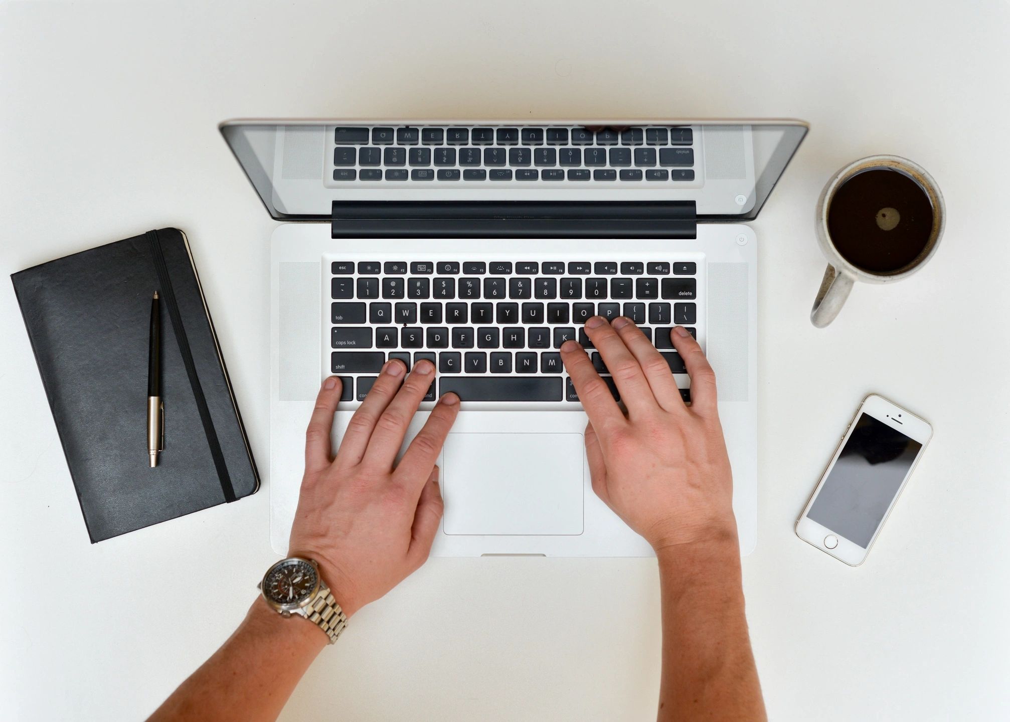 Hands typing on a laptop keyboard, with a notebook, smartphone, and cup of coffee nearby on a white desk.