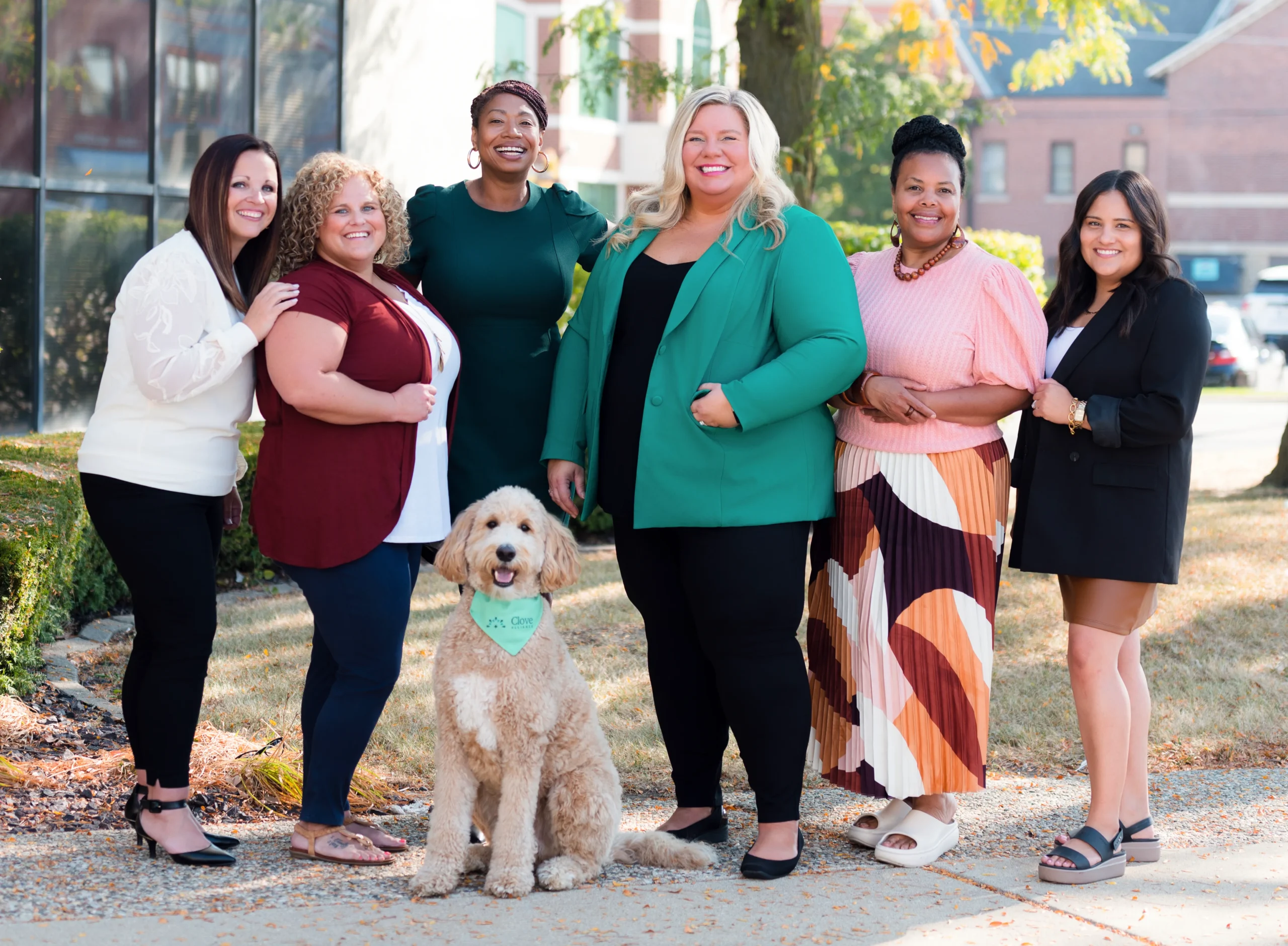 A group of six women who form Clove Alliance's leadership team, and a dog stand outdoors, smiling at the camera. Trees and buildings are in the background.