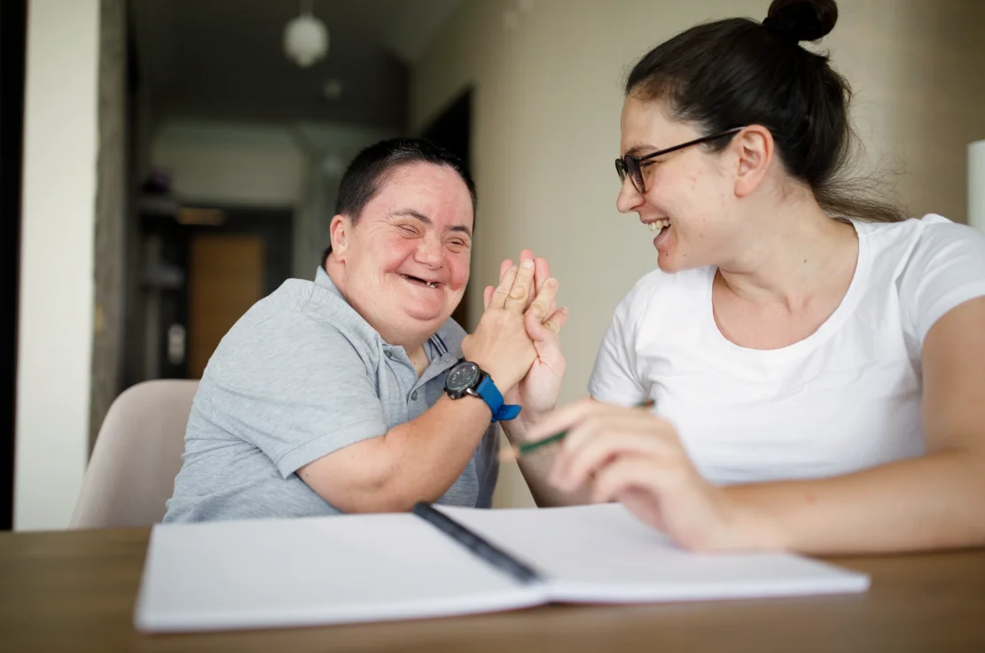 A person with Down's syndrome short hair and a gray shirt smiles and high-fives a person with glasses and a white shirt. A notebook and pen are on the table in front of them.