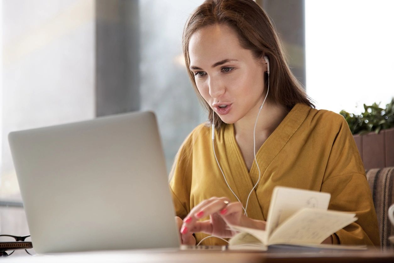 Person wearing earphones sits at a laptop with an open notebook, appearing focused.