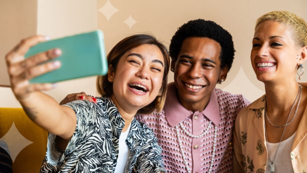 Three queer friends smile as one holds out a smartphone to take a selfie photo of the group.