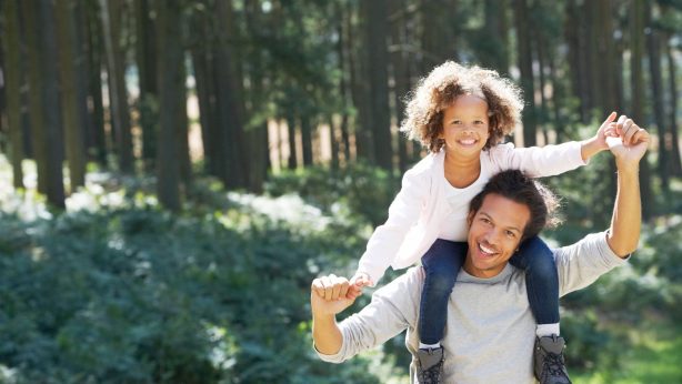 A man carries a smiling child on his shoulders while walking in a sunlit forest. Both are holding hands and smiling.