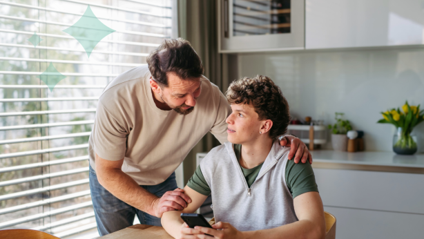 A father stands beside a seated teenage son holding a phone at a table.