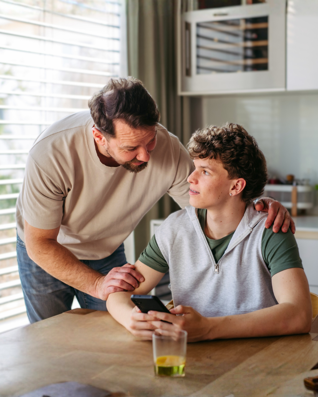 A father stands beside a seated teenage son holding a phone at a table.