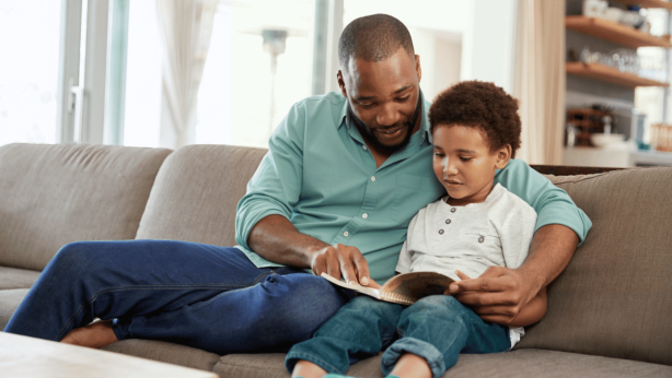 An father and a child sit together on a couch, reading a book. The adult is pointing at the page, and both look focused on the book.